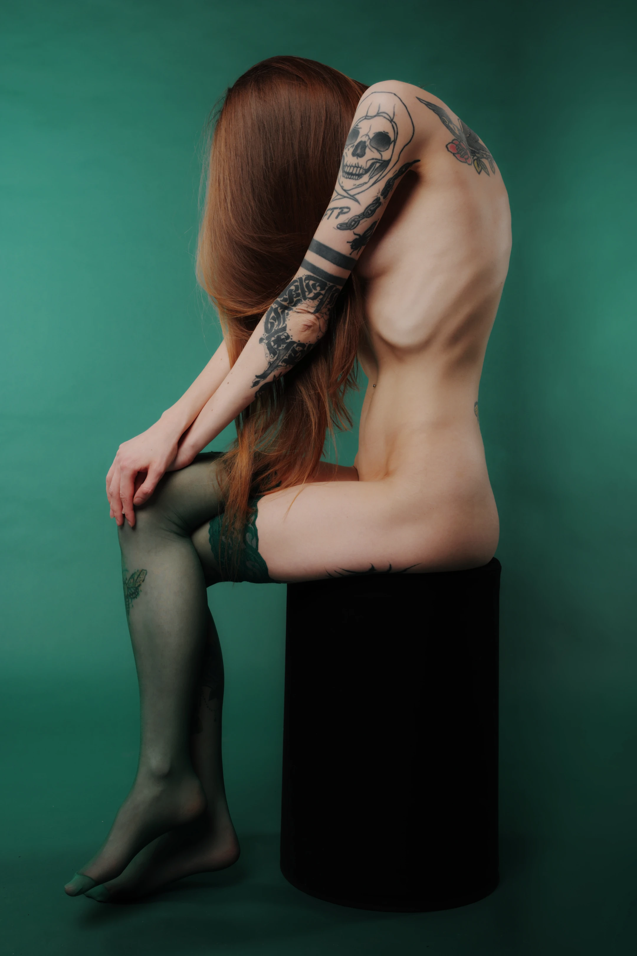 A nude woman with long, flowing auburn hair sits on a black cylindrical stool against a solid, deep emerald-green backdrop, creating a moody, surreal atmosphere. Her body is mostly bare, revealing intricate black ink tattoos covering her arms, shoulders, and legs, including a prominent skull design on her upper arm and tribal patterns along her forearm. She wears sheer black tights with subtle lace trim at the thigh, emphasizing her tattooed skin and the stark contrast with the dark stool and vibrant background. Her pose is introspective: she leans forward slightly, one arm draped over her knee, the other resting near her thigh.